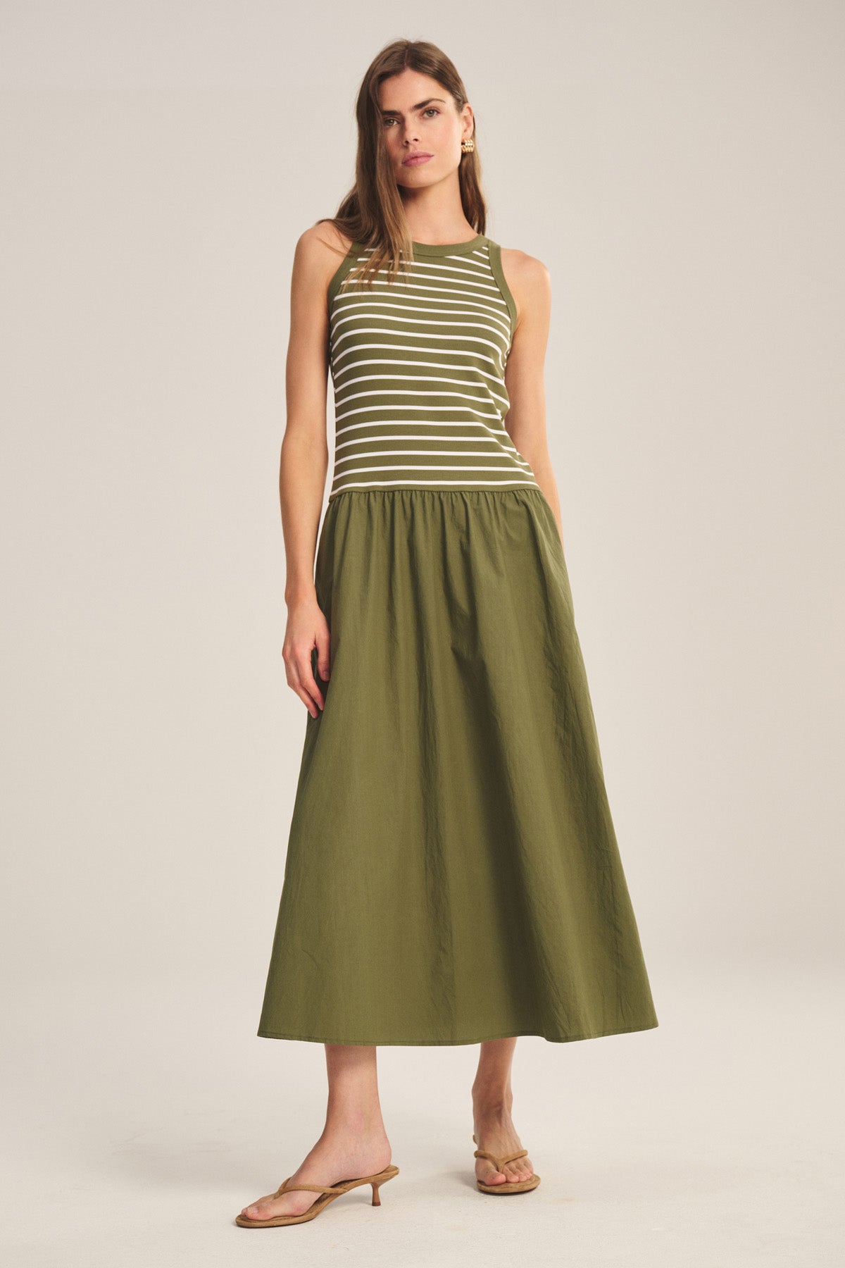 A woman wears the ROMINA DRESS by Velvet by Graham & Spencer—an olive green and white striped, sleeveless dress—paired with tan heeled sandals, standing against a plain background.-41338484916417