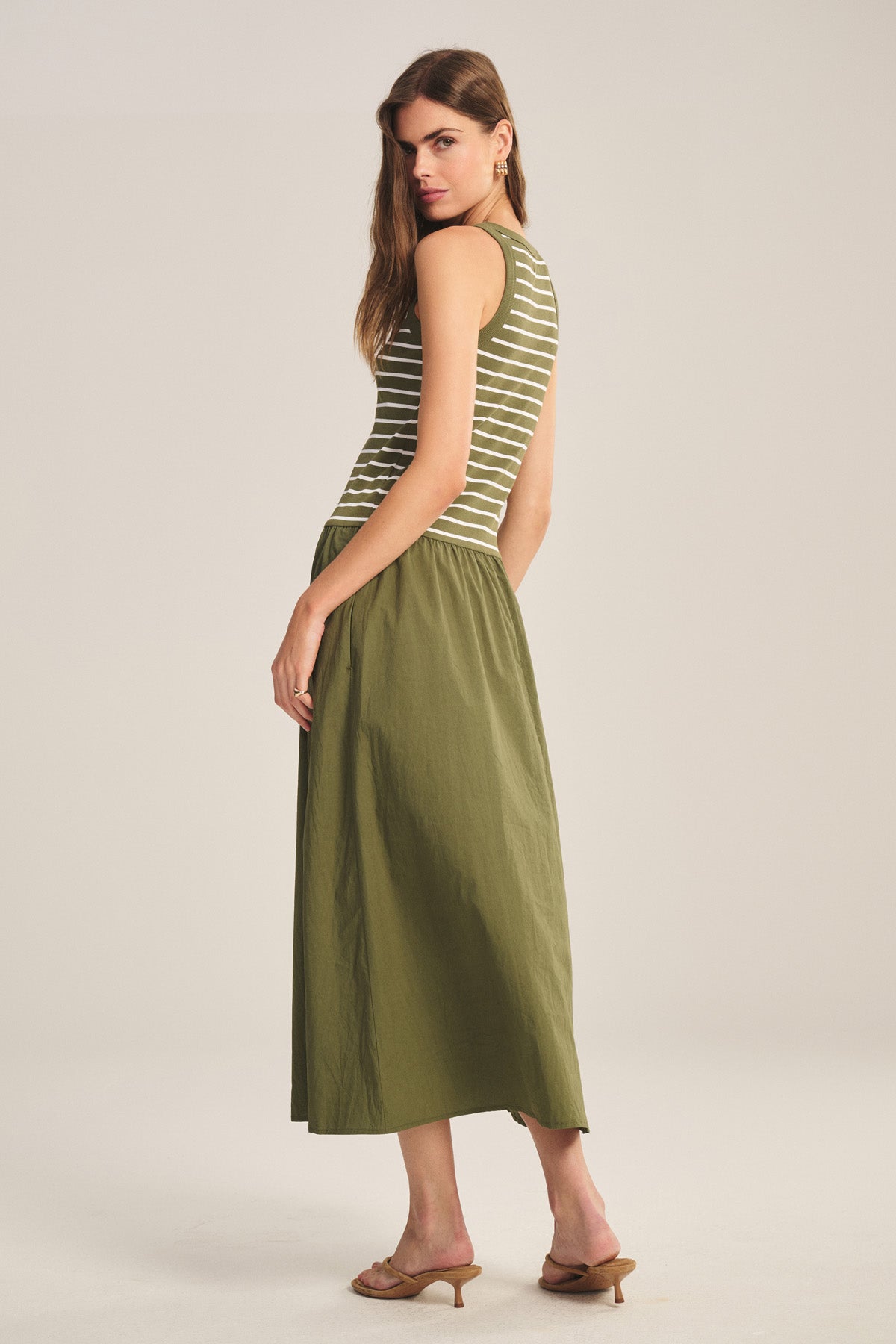 A woman stands in a studio wearing the ROMINA DRESS by Velvet by Graham & Spencer, a sleeveless green and white striped dress, paired with tan heeled sandals. She is turned slightly to the side.-41338484883649