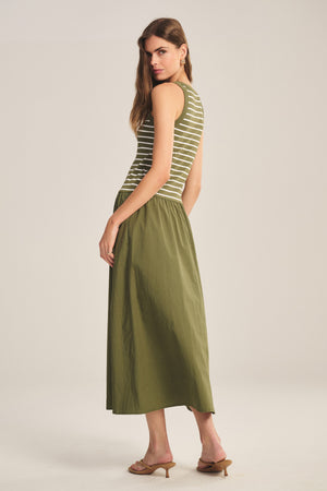 A woman stands in a studio wearing the ROMINA DRESS by Velvet by Graham & Spencer, a sleeveless green and white striped dress, paired with tan heeled sandals. She is turned slightly to the side.