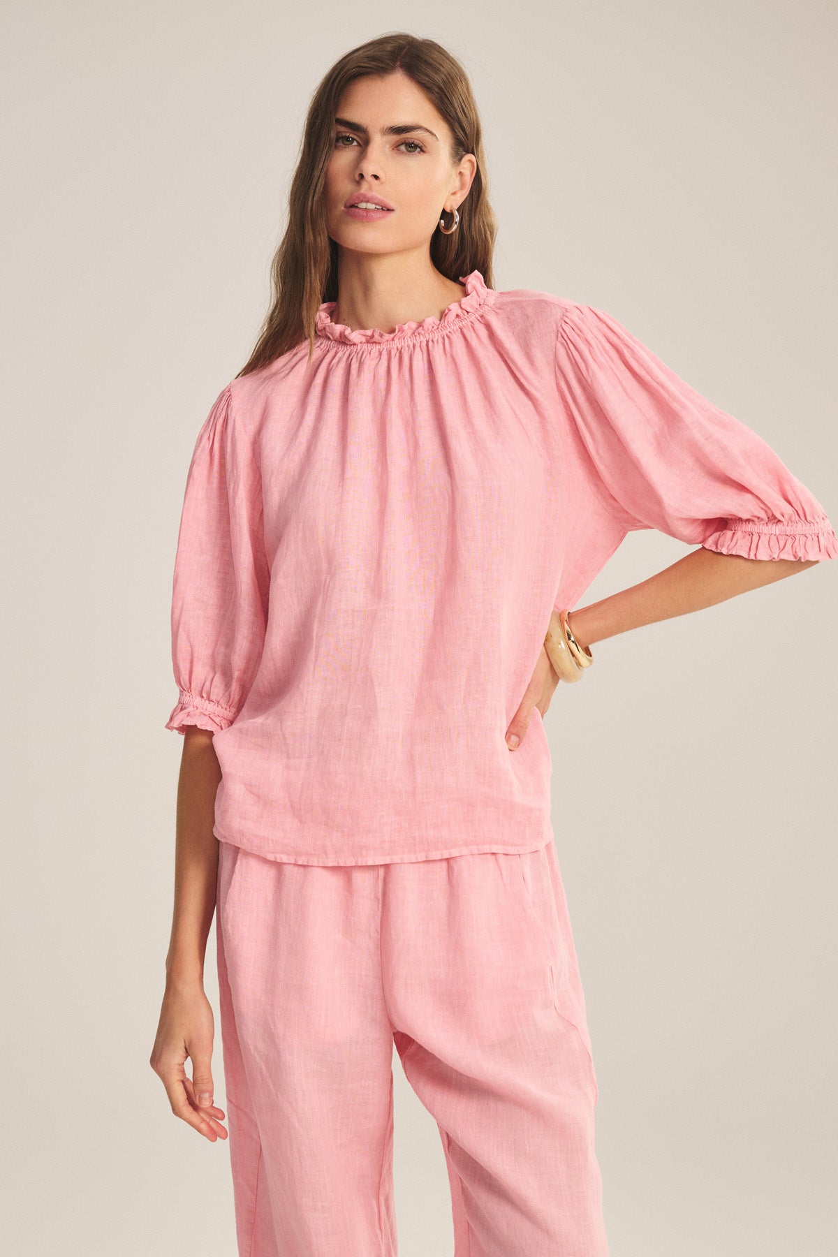A woman wears the ROSABEL LINEN TOP by Velvet by Graham & Spencer—a loose, light pink, long-sleeve blouse with ruffled collar and cuffs—paired with matching pants, standing against a neutral background.-41296981164225