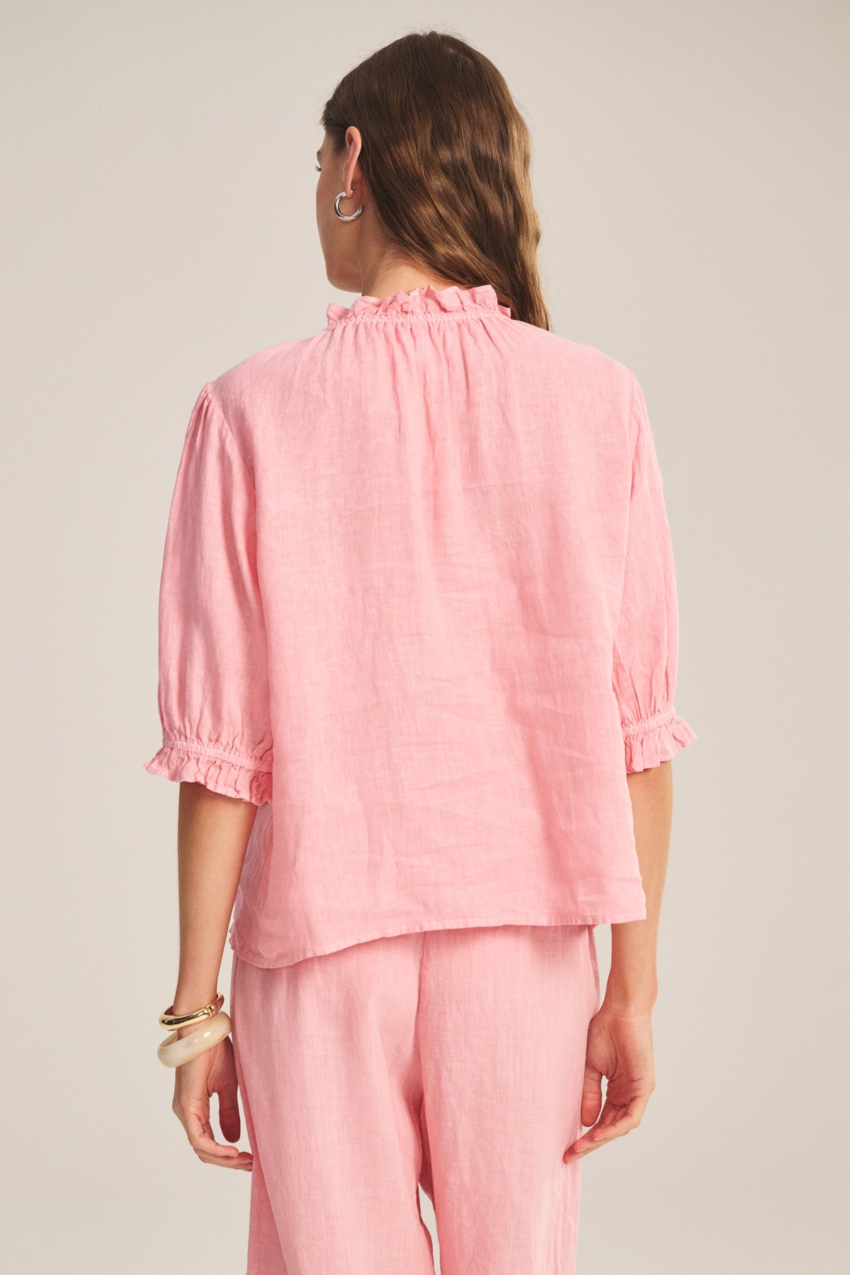 A person stands facing away, wearing the ROSABEL LINEN TOP by Velvet by Graham & Spencer—a light pink, long-sleeve blouse with ruffled collar and cuffs—paired with matching pink pants.