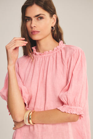 A woman wears the ROSABEL LINEN TOP by Velvet by Graham & Spencer—a loose-fitting pink blouse with ruffled sleeves and collar—paired with gold hoop earrings and cream bangles, standing against a neutral background.
