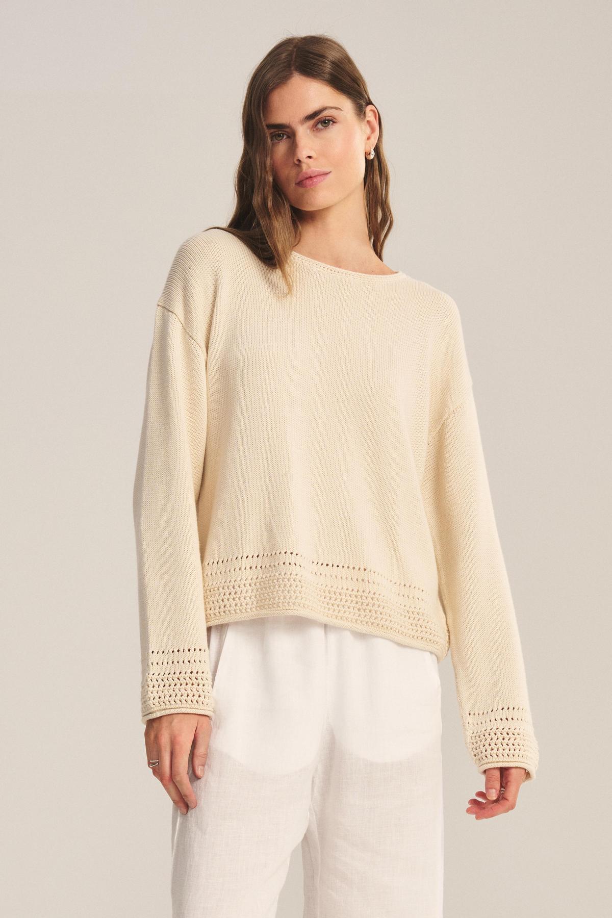 A woman wears the ROSALYN SWEATER by Velvet by Graham & Spencer—a loose, cream knit in soft Pima cotton with crocheted hem and cuffs—paired with white pants against a plain background.