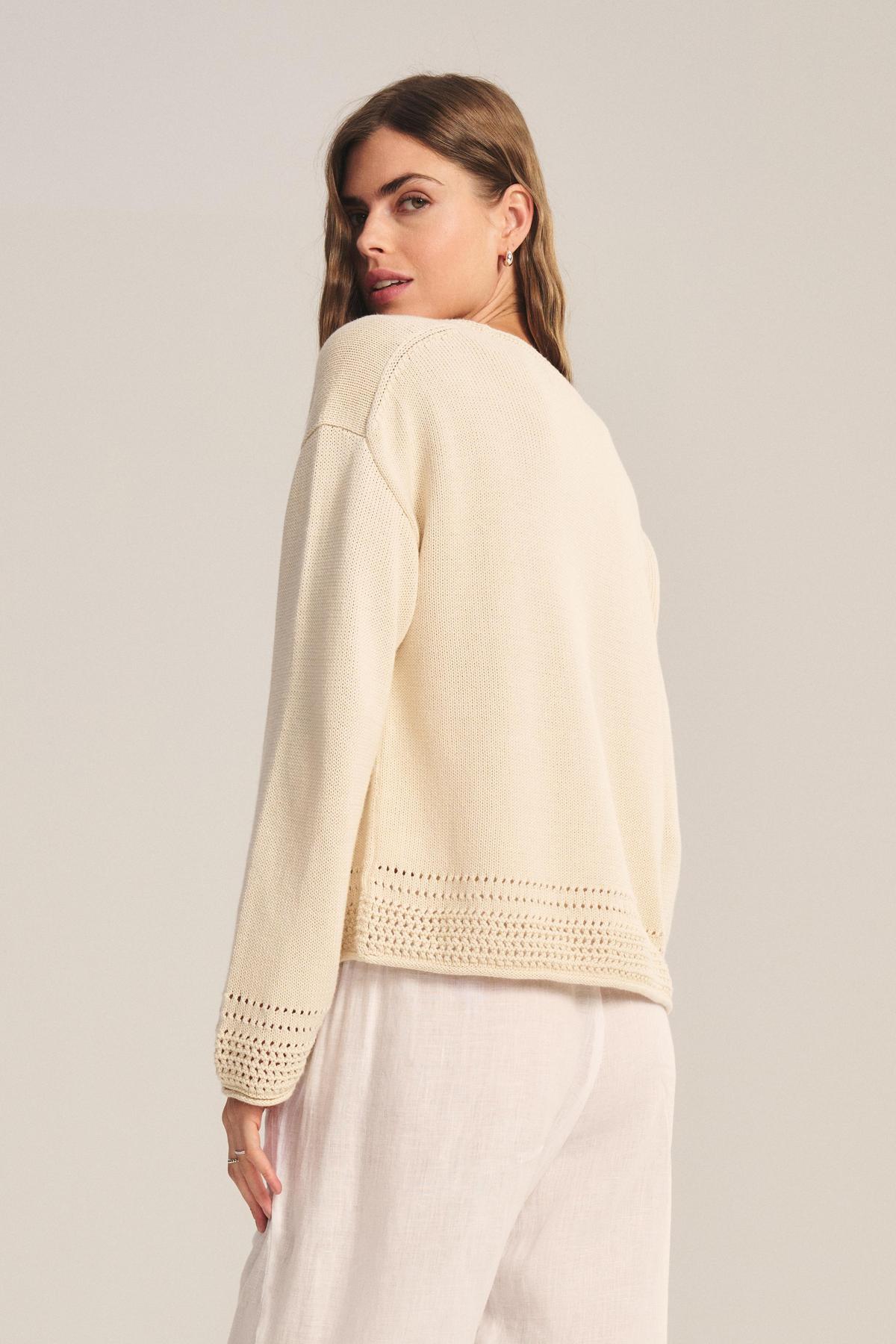 A woman stands sideways against a plain background, wearing the ROSALYN SWEATER by Velvet by Graham & Spencer—a cream Pima cotton knit with crocheted details at the hem and cuffs—paired with loose white pants.