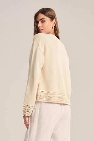 A woman stands sideways against a plain background, wearing the ROSALYN SWEATER by Velvet by Graham & Spencer—a cream Pima cotton knit with crocheted details at the hem and cuffs—paired with loose white pants.