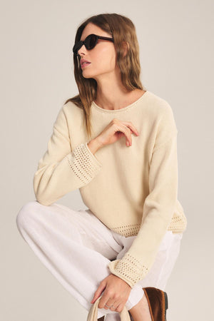 A woman in sunglasses, white pants, and the ROSALYN SWEATER by Velvet by Graham & Spencer crouches with a tote bag against a plain background.