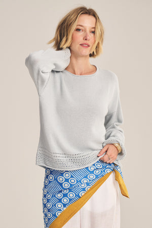 A woman models the Velvet by Graham & Spencer ROSALYN SWEATER in light gray Pima cotton with subtle pointelle detailing, paired with a blue, white, and yellow patterned wrap skirt, set against a neutral background.