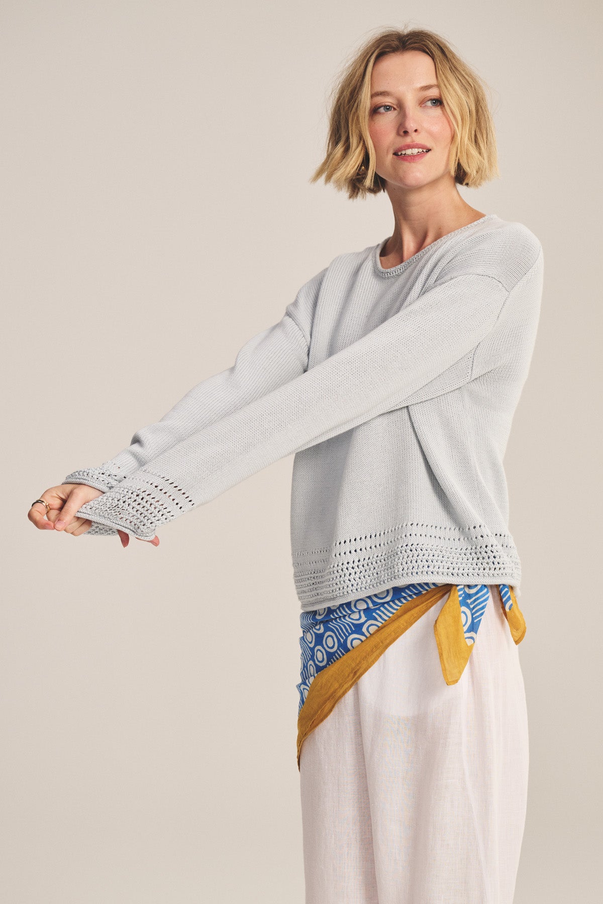 A woman with short blonde hair wears the Velvet by Graham & Spencer ROSALYN SWEATER in light gray Pima cotton knit with pointelle details and a relaxed drop-shoulder fit, styled with loose white pants and a blue and yellow waist scarf.-40909979549889