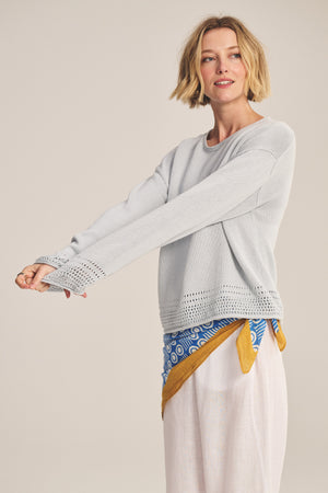 A woman with short blonde hair wears the Velvet by Graham & Spencer ROSALYN SWEATER in light gray Pima cotton knit with pointelle details and a relaxed drop-shoulder fit, styled with loose white pants and a blue and yellow waist scarf.