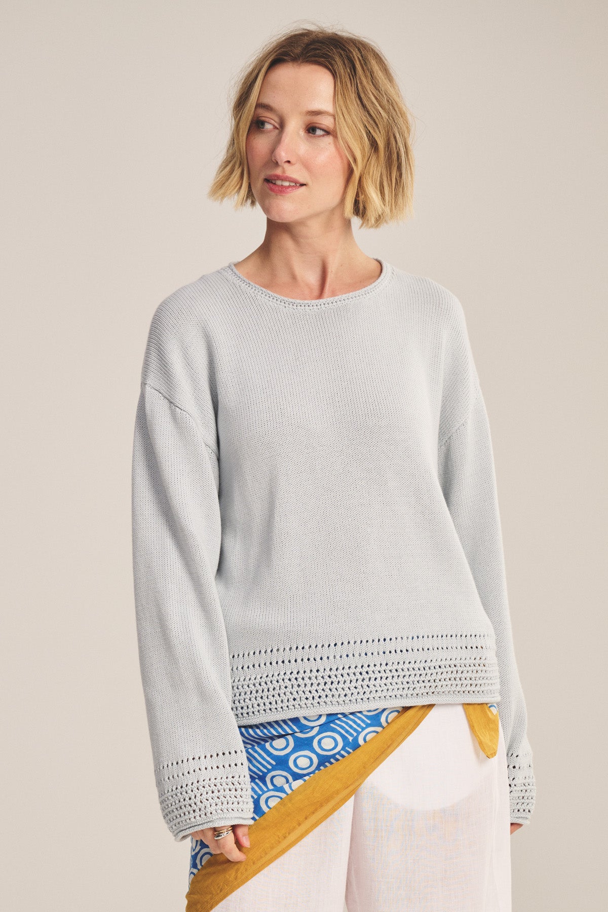 A woman with short blonde hair wears the ROSALYN SWEATER by Velvet by Graham & Spencer, a light gray knit with pointelle detailing, paired with a blue and white patterned skirt and yellow sash against a neutral background.-40909978763457
