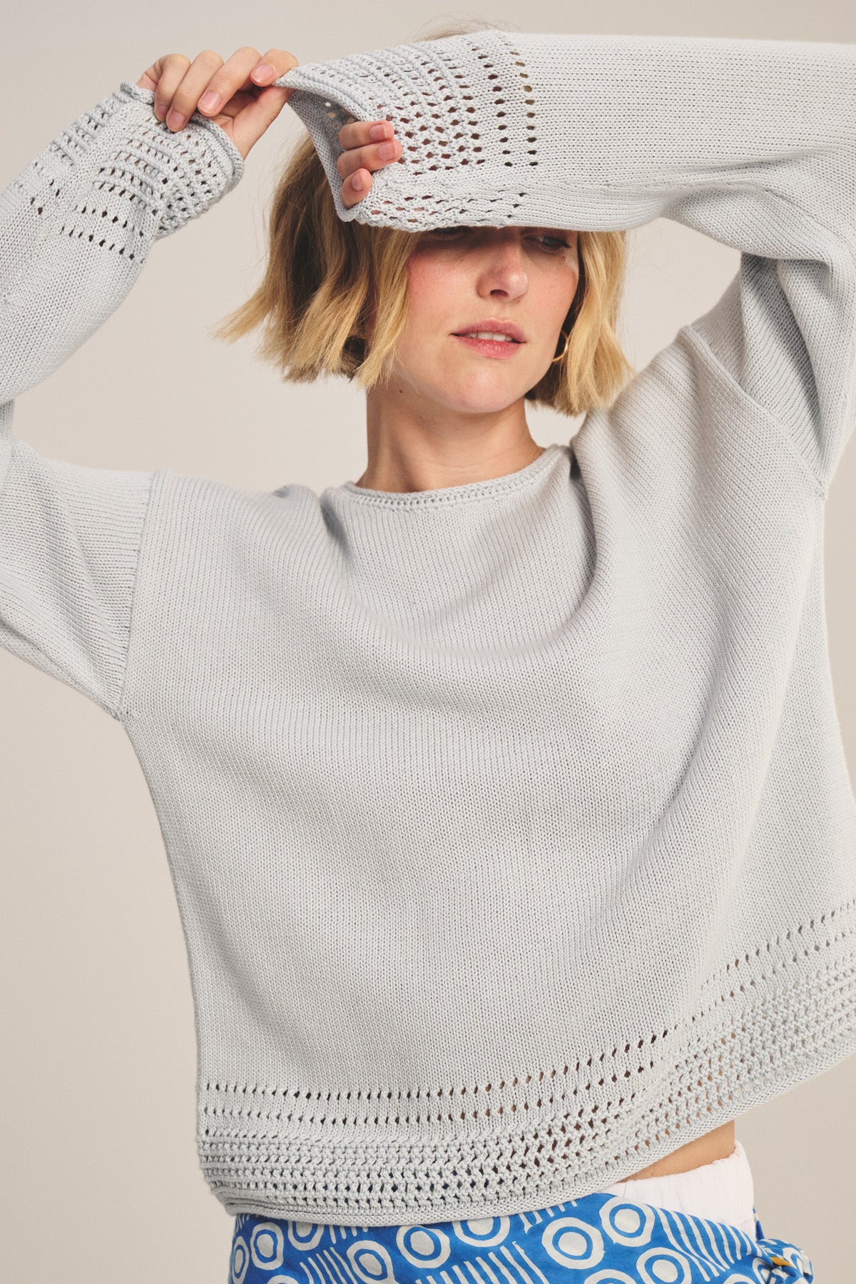 A woman wears the Velvet by Graham & Spencer ROSALYN SWEATER in light gray Pima cotton with pointelle details, paired with blue and white patterned pants, holding one arm across her face and smiling slightly.-40909978697921