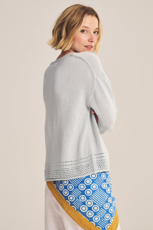 A woman with short blonde hair wears the Velvet by Graham & Spencer ROSALYN SWEATER in light gray Pima cotton, featuring a relaxed drop-shoulder fit, and a white skirt with a blue geometric and yellow border pattern, back slightly turned.