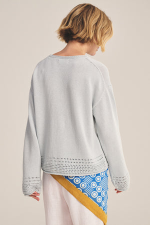 A person with short blond hair, seen from behind, wears the ROSALYN SWEATER by Velvet by Graham & Spencer in light gray Pima cotton with a relaxed drop-shoulder fit, paired with a patterned blue, white, and yellow skirt.