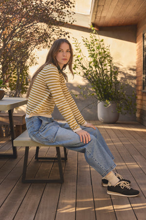 A woman wearing the ROXY SWEATER by Velvet by Graham & Spencer and blue jeans sits on a bench on a wooden patio, with plants and sunlight in the background.