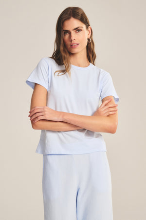 A woman stands with arms crossed, wearing the Velvet by Graham & Spencer RYLAND TEE—a light blue cotton jersey T-shirt with a boxy silhouette—paired with matching pants against a plain background.
