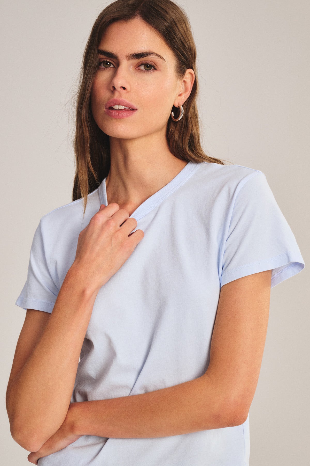 Woman with long brown hair wears the RYLAND TEE by Velvet by Graham & Spencer—a light blue cotton jersey crew neck—standing against a neutral background and looking at the camera.-41044143833281