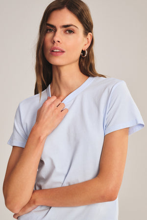 Woman with long brown hair wears the RYLAND TEE by Velvet by Graham & Spencer—a light blue cotton jersey crew neck—standing against a neutral background and looking at the camera.