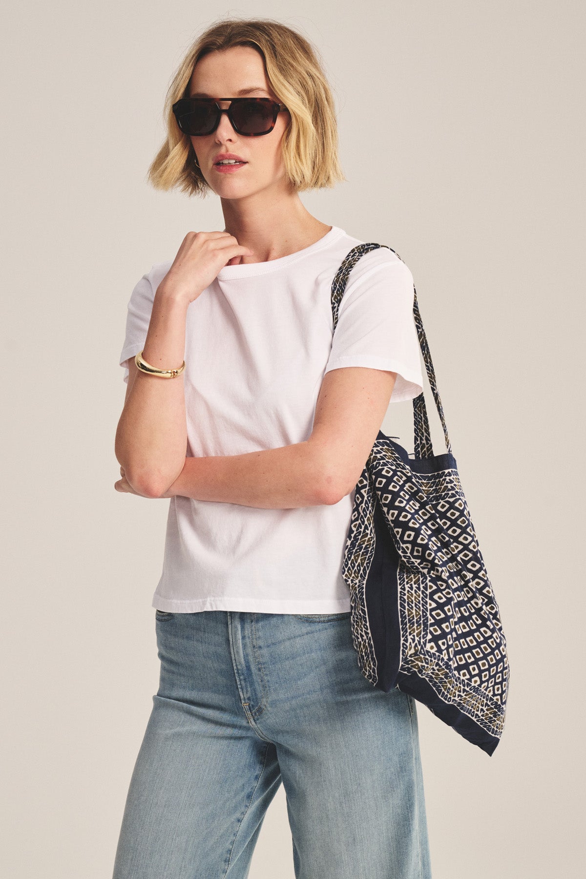 A woman in sunglasses, light blue jeans, and the RYLAND TEE by Velvet by Graham & Spencer—a white cotton jersey crewneck with a boxy fit—holds a patterned tote bag over her shoulder against a plain background.-41178051903681