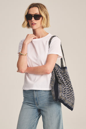 A woman in sunglasses, light blue jeans, and the RYLAND TEE by Velvet by Graham & Spencer—a white cotton jersey crewneck with a boxy fit—holds a patterned tote bag over her shoulder against a plain background.