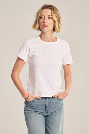 A woman stands with her hands in her pockets against a neutral background, wearing the RYLAND TEE by Velvet by Graham & Spencer and blue jeans.