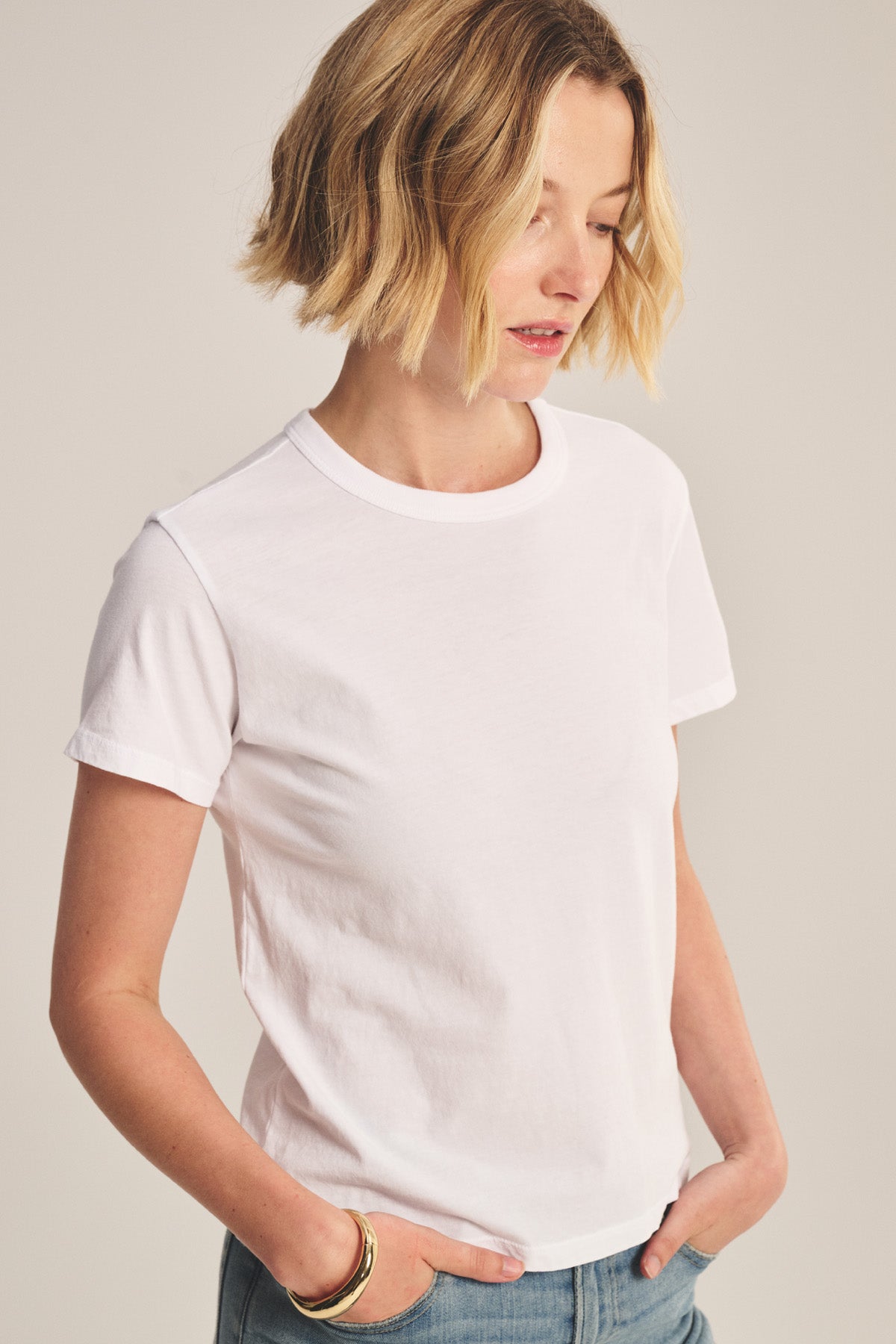 A woman with short blonde hair wears the RYLAND TEE by Velvet by Graham & Spencer—a plain white, boxy silhouette top—paired with blue jeans, standing with hands in her pockets and looking down.