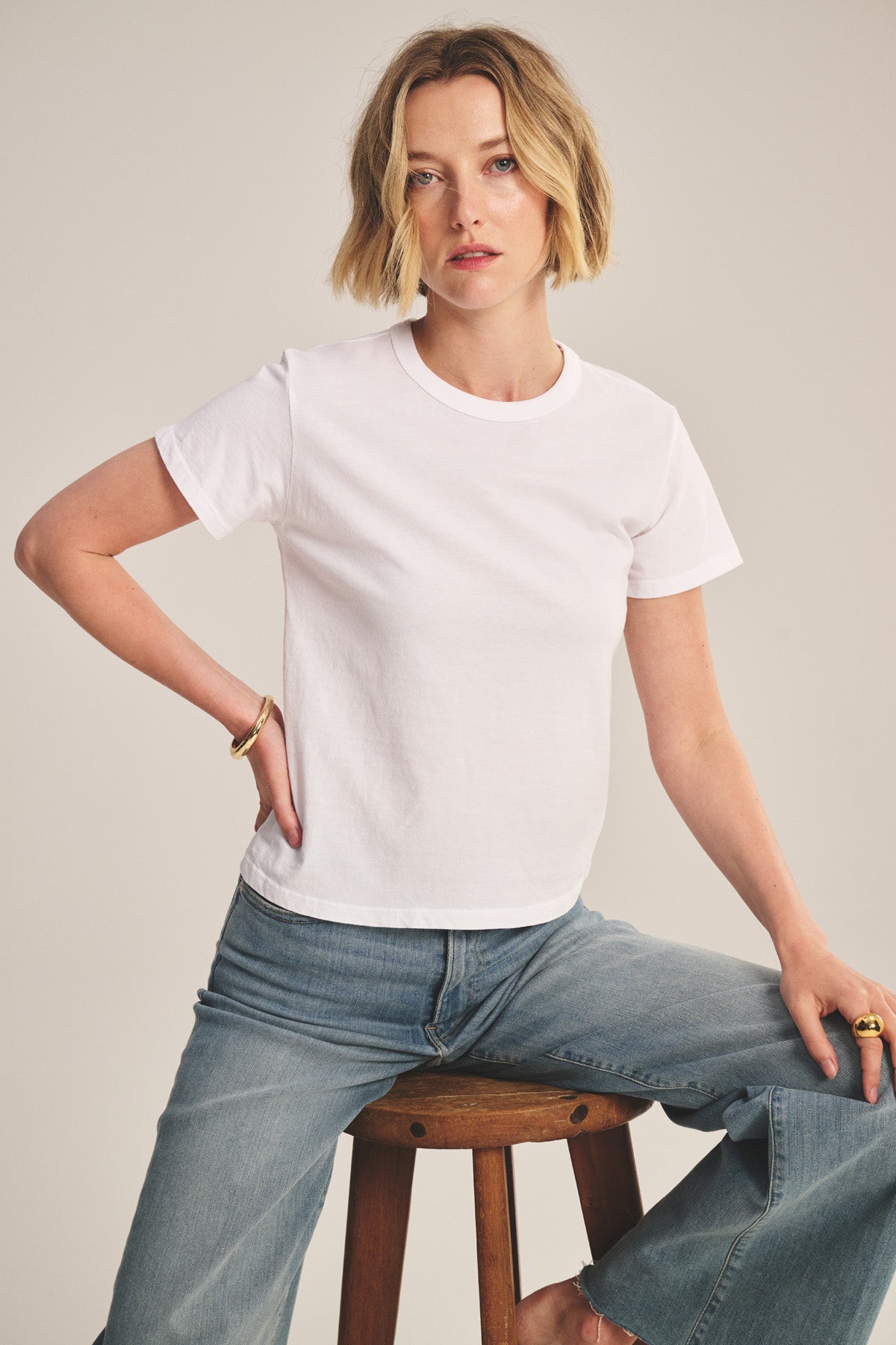 A person with short blonde hair wears the Velvet by Graham & Spencer RYLAND TEE and blue jeans while sitting on a wooden stool against a neutral background.-41178051838145