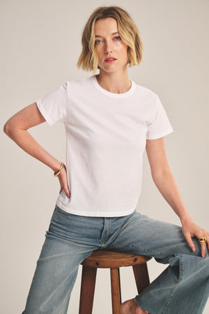 A person with short blonde hair wears the Velvet by Graham & Spencer RYLAND TEE and blue jeans while sitting on a wooden stool against a neutral background.