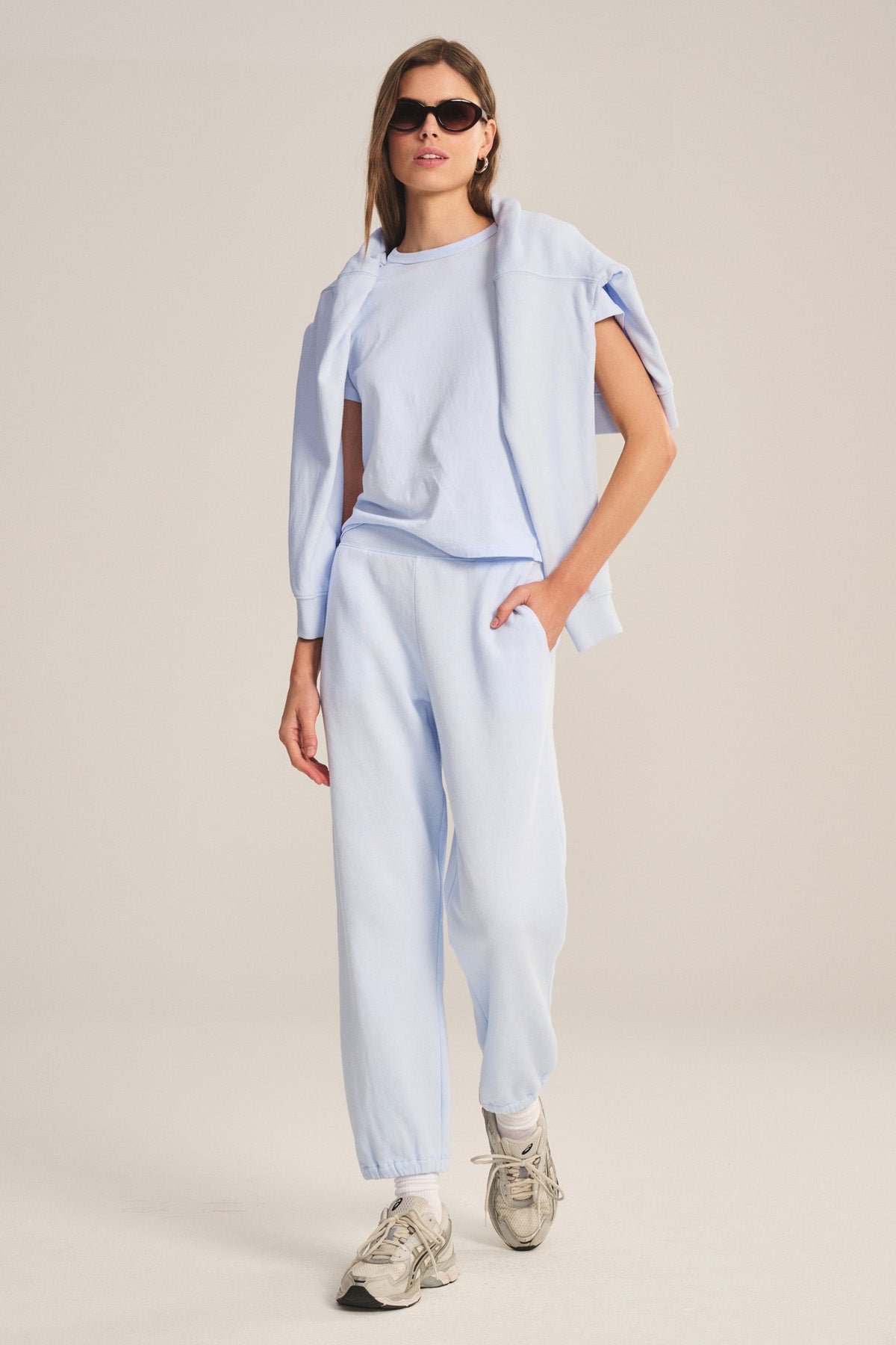 A person stands in a neutral studio wearing light blue ZARA SWEATPANT by Velvet by Graham & Spencer, matching joggers, sneakers, a relaxed fit sweatshirt draped over their shoulders, and sunglasses.-41044143800513