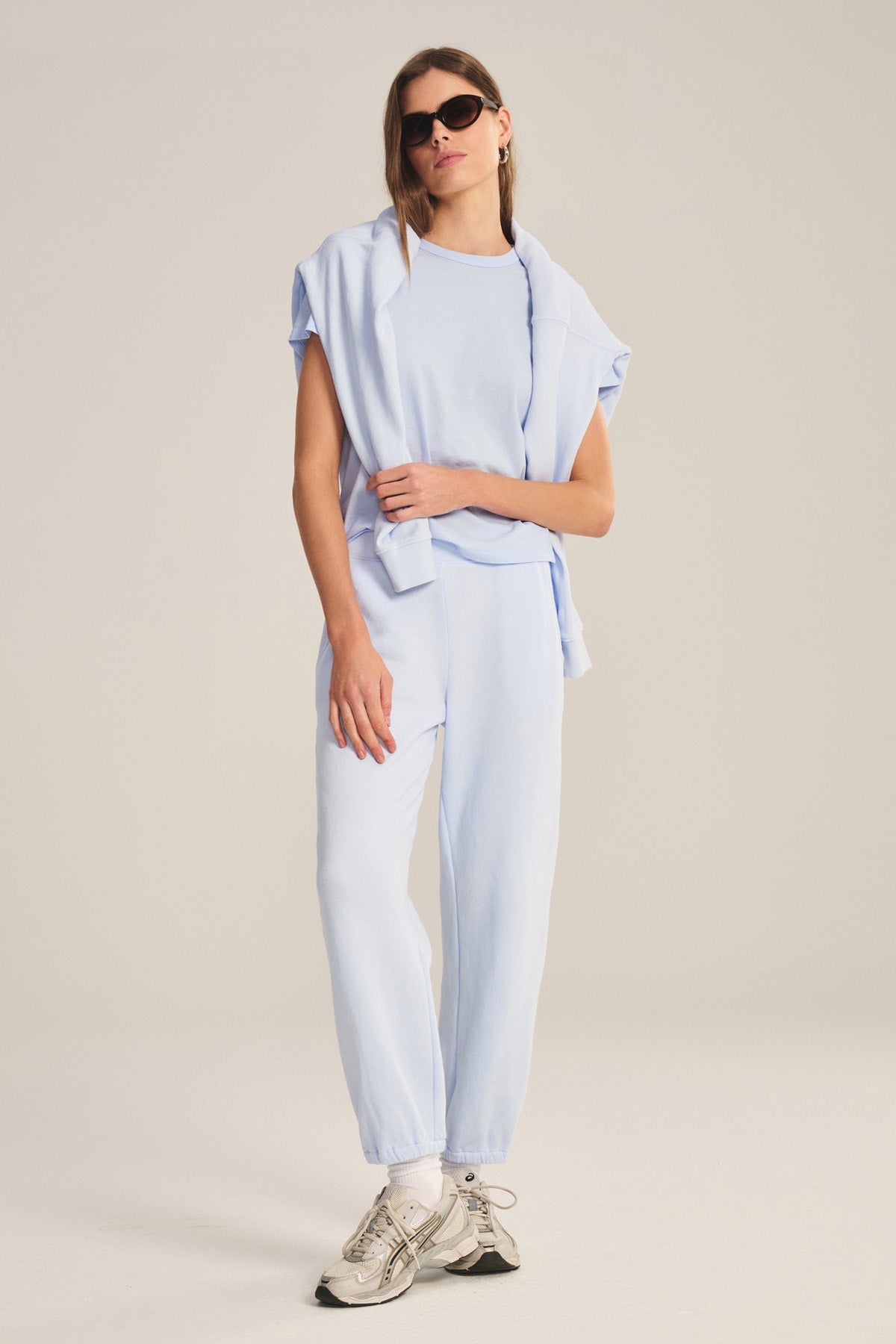 A woman wears the RYLAND TEE by Velvet by Graham & Spencer—a light blue cotton jersey top paired with matching sweatpants, a sweatshirt draped over her shoulders, sunglasses, white socks, and sneakers.-41044143767745