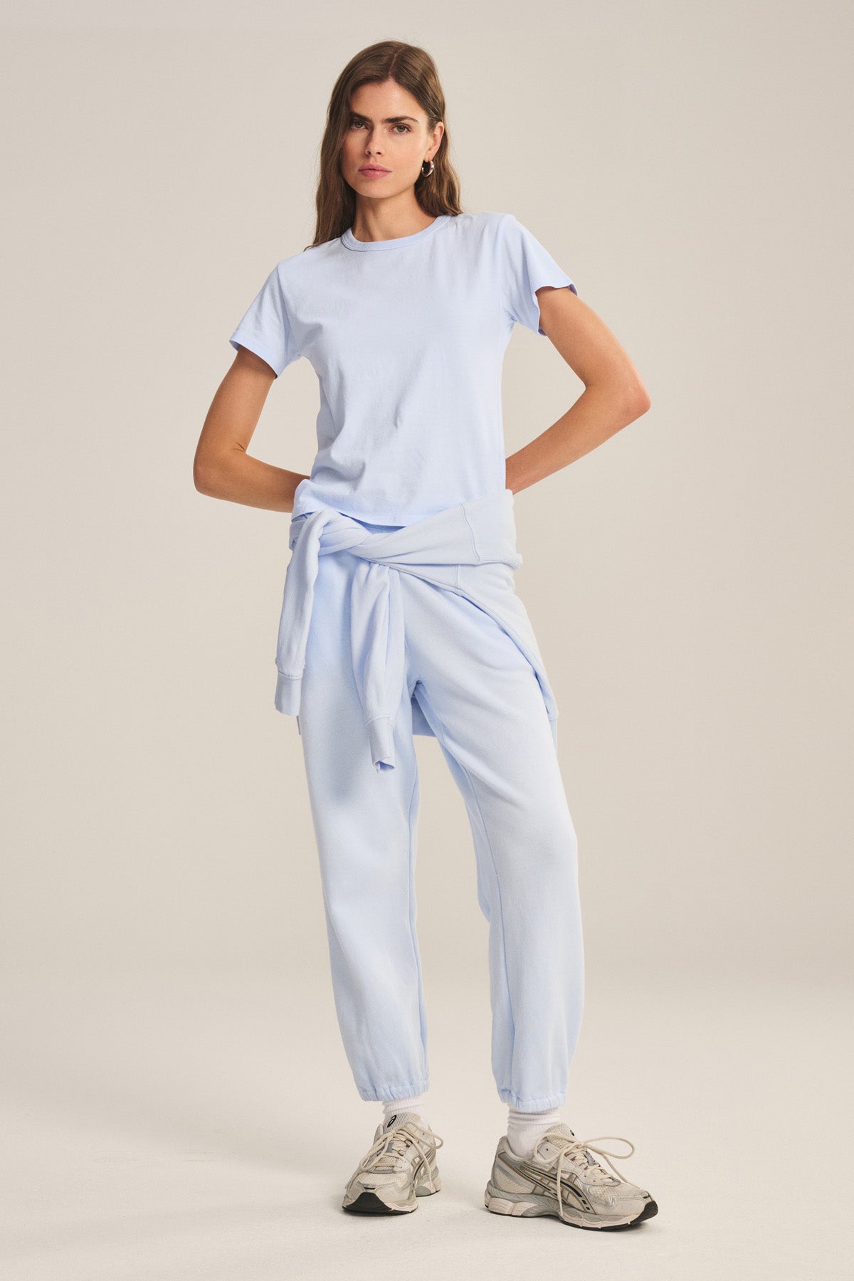 A woman wears a light blue t-shirt and Velvet by Graham & Spencer ZARA SWEATPANT in cotton fleece, with a matching sweatshirt tied at her waist and gray athletic shoes.