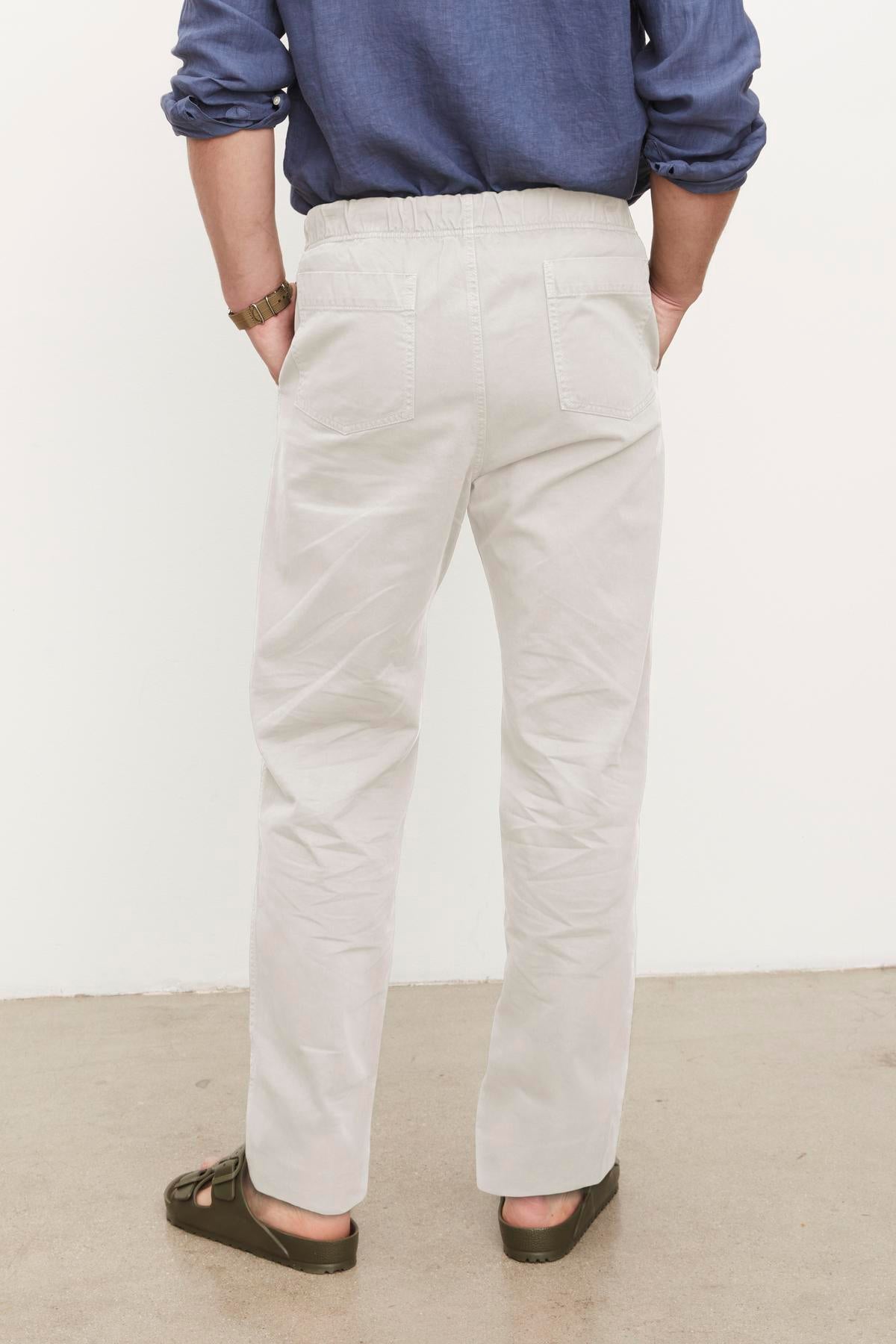 A person standing with their back to the camera, wearing light-colored cotton sanded twill Branson Pants by Velvet by Graham & Spencer and a blue shirt with rolled-up sleeves.-36329591144641