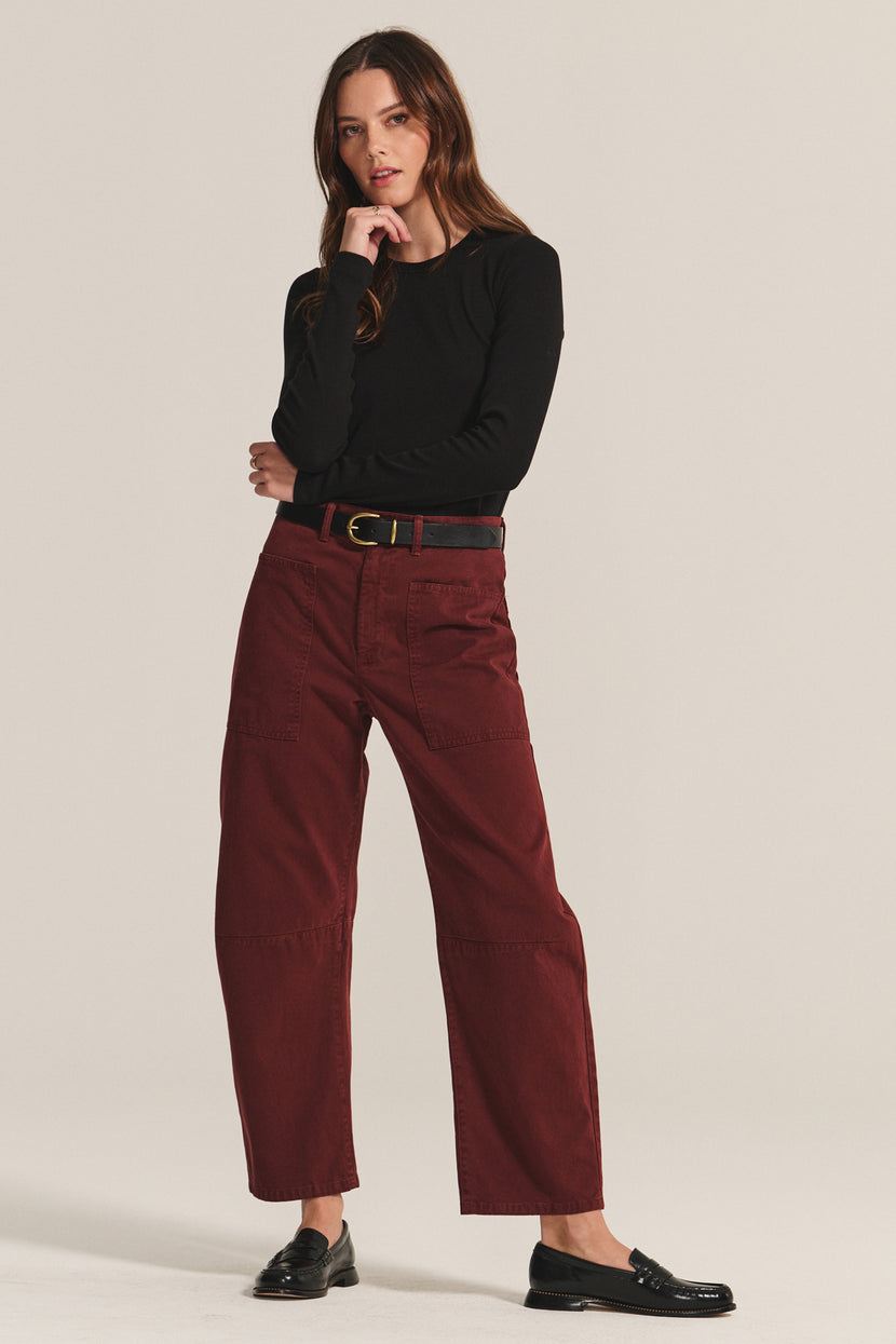 A woman stands against a plain background, wearing a black long-sleeve top, Velvet by Graham & Spencer's BRYLIE SANDED TWILL UTILITY PANT in maroon with patch pockets and a black belt, finished with black loafers.