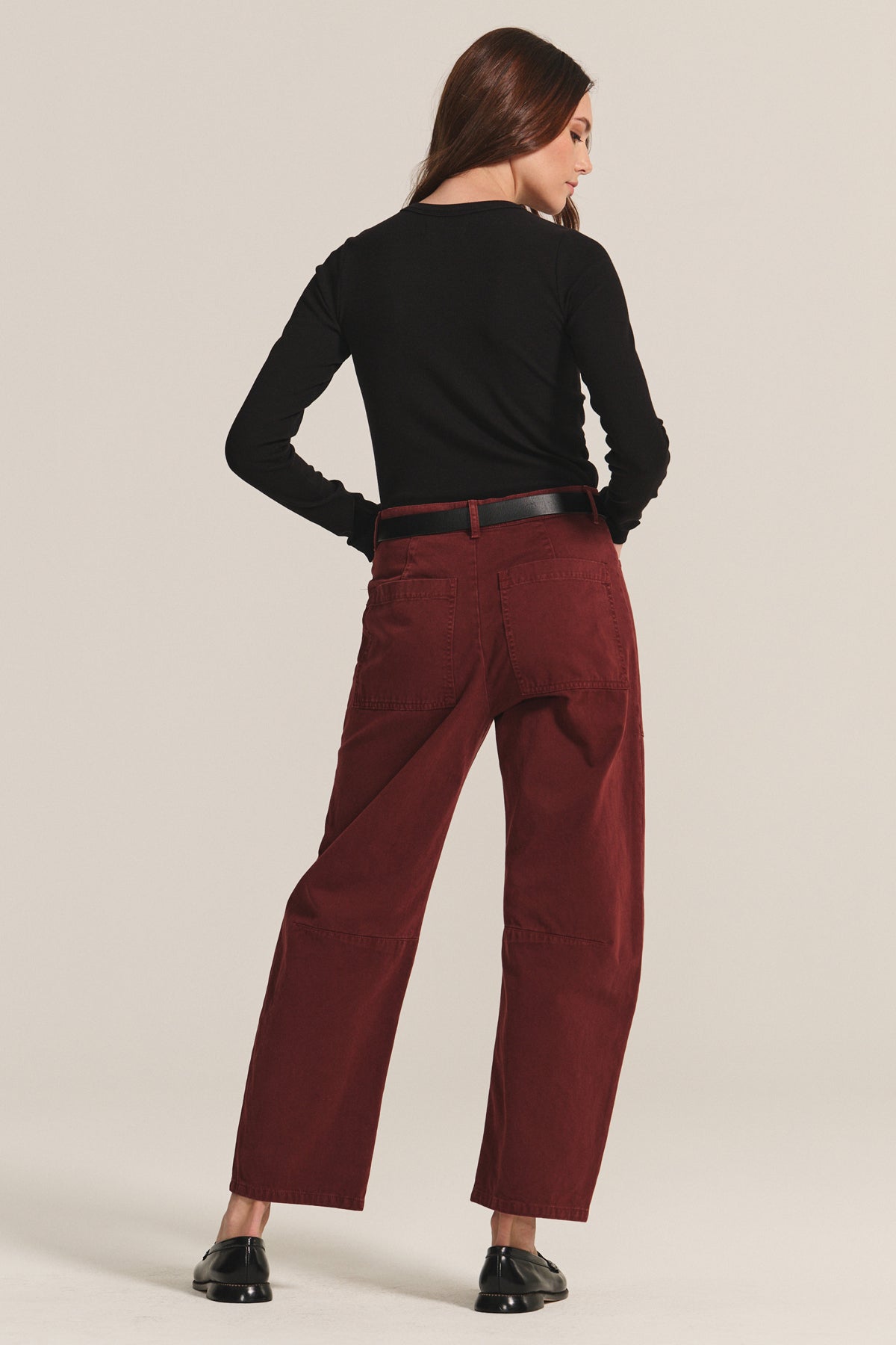   A person stands facing away, wearing a black long-sleeve top, black belt, black shoes, and Velvet by Graham & Spencer's BRYLIE SANDED TWILL UTILITY PANT in maroon against a plain background. 