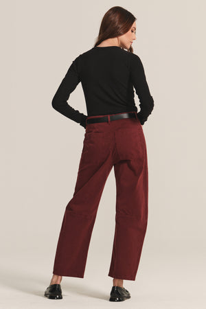 A person stands facing away, wearing a black long-sleeve top, black belt, black shoes, and Velvet by Graham & Spencer's BRYLIE SANDED TWILL UTILITY PANT in maroon against a plain background.