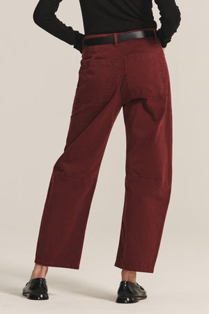 Person wears Velvet by Graham & Spencer's BRYLIE SANDED TWILL UTILITY PANT—high-waisted, loose maroon trousers with patch pockets—paired with a black belt, black long-sleeve top, and black shoes, shown from the back.