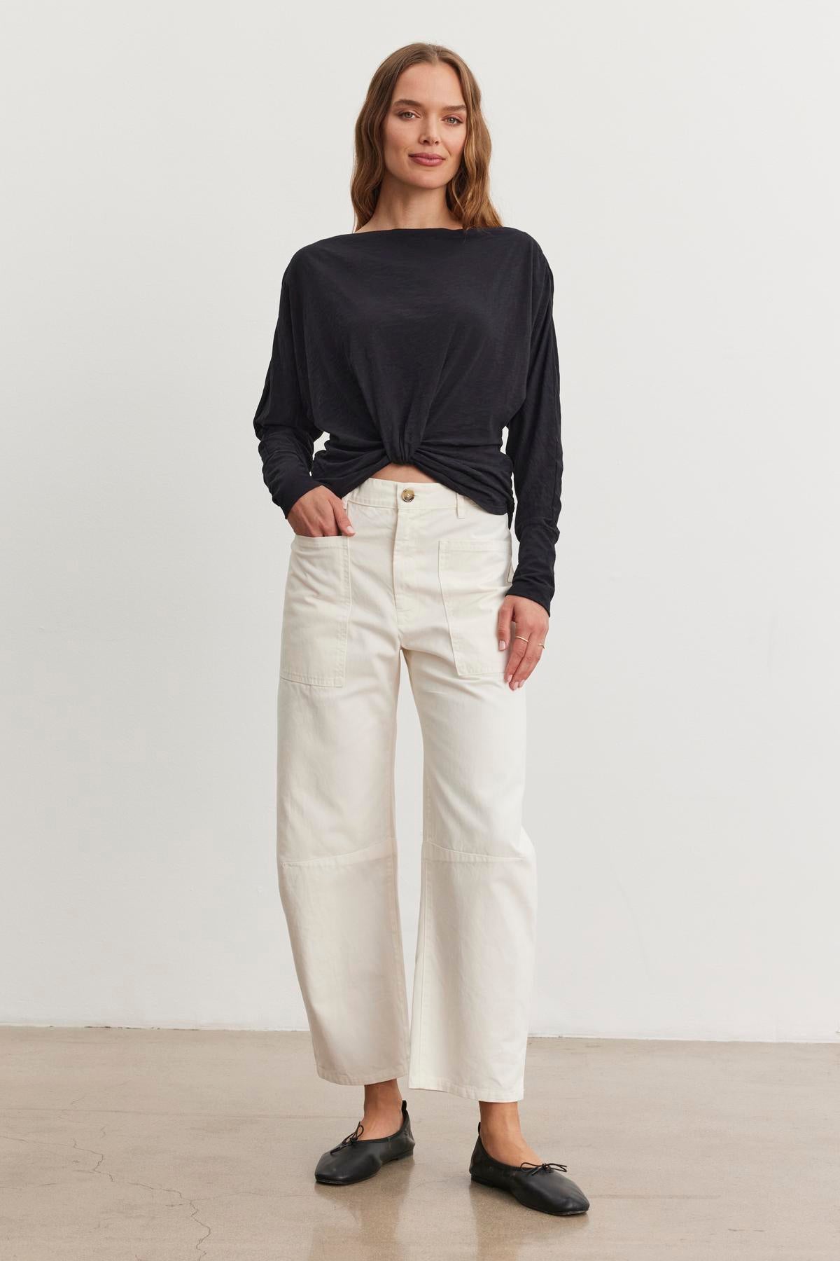   A person stands against a plain background wearing a black long-sleeve top, BRYLIE SANDED TWILL UTILITY PANT by Velvet by Graham & Spencer in white cotton, and black shoes. 