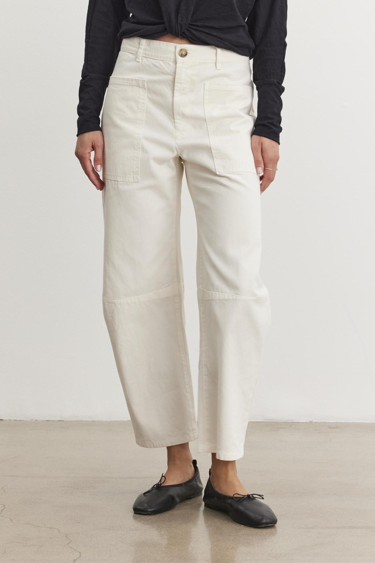 A person stands on a polished concrete floor, wearing Velvet by Graham & Spencer's cream BRYLIE SANDED TWILL UTILITY PANT and a black long-sleeve top. Black ballet flats complete the casual look.-38512513745089