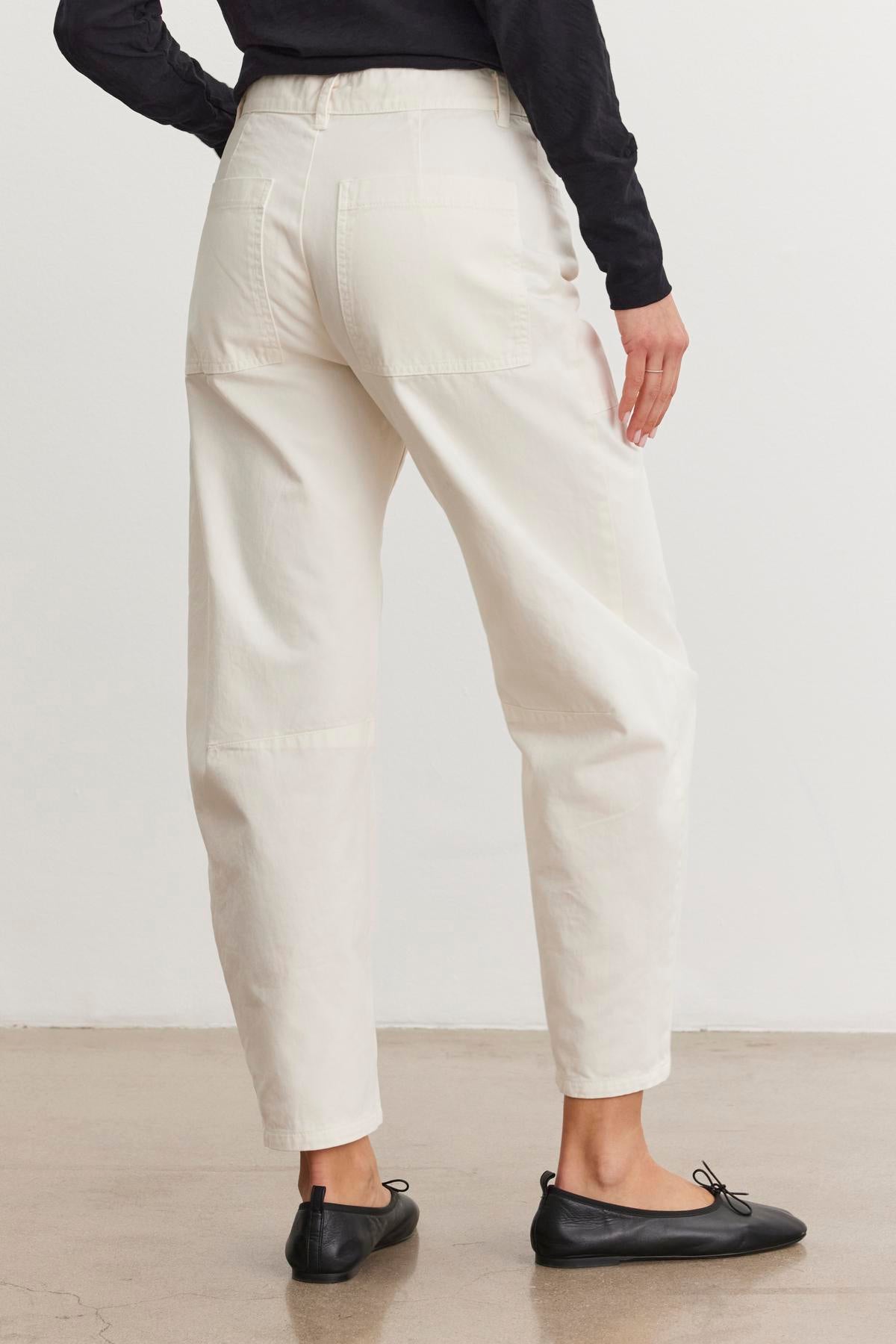 A person wearing Velvet by Graham & Spencer's Brylie Sanded Twill Utility Pant with patch pockets and black flats is viewed from behind, standing on a concrete floor.-38512513843393