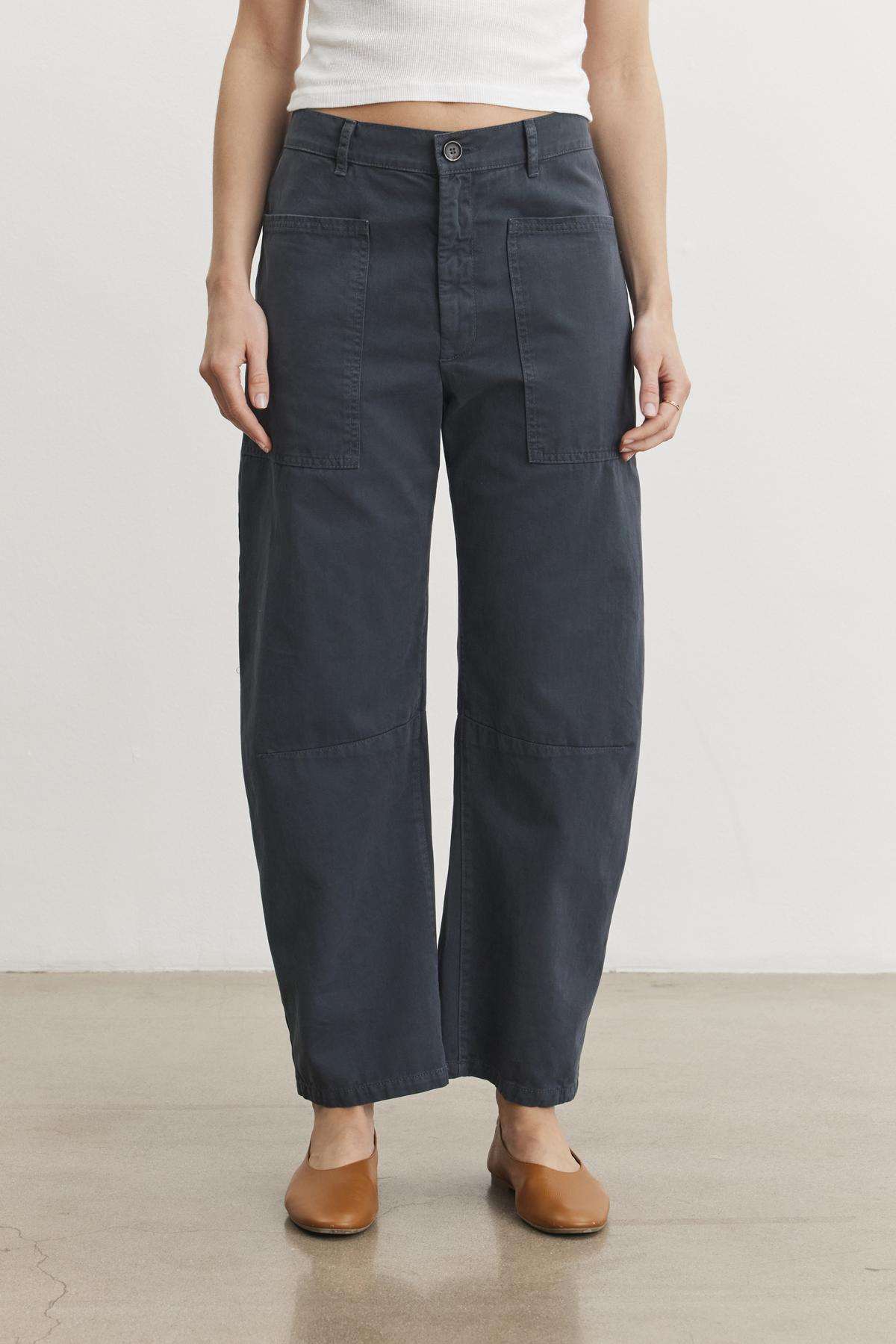   The person is sporting Velvet by Graham & Spencer's BRYLIE SANDED TWILL UTILITY PANT in dark blue with a white top, standing on a polished floor. 