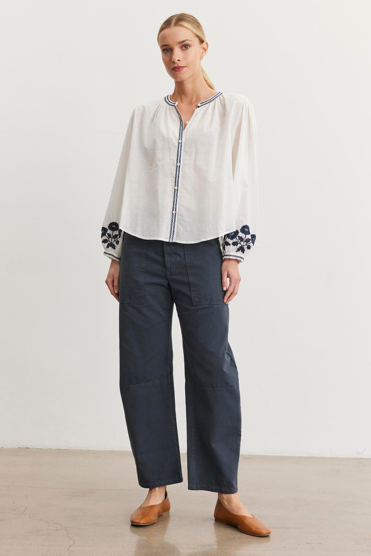   A person stands against a plain white background, wearing a white blouse with floral sleeves, Velvet by Graham & Spencer's dark BRYLIE SANDED TWILL UTILITY PANT featuring patch pockets, and brown shoes. 