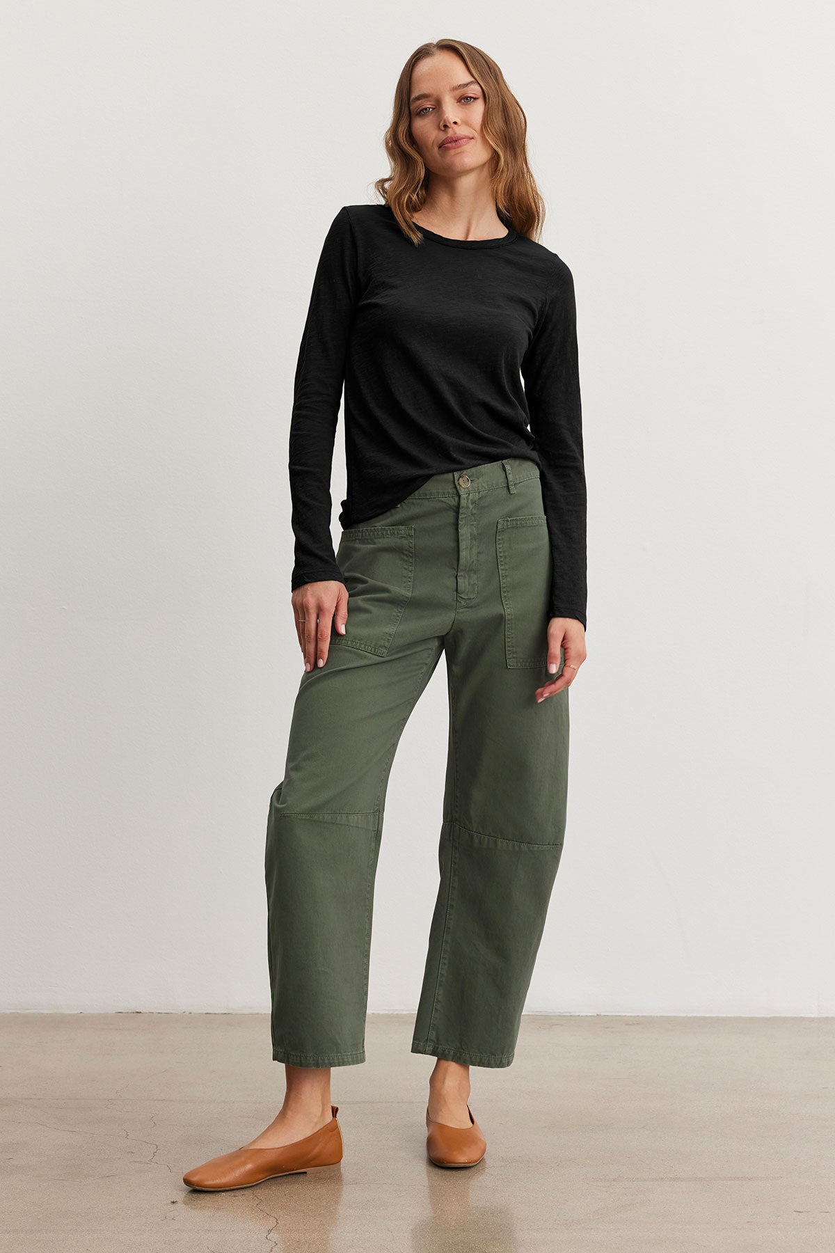   A person stands against a plain background wearing a black long-sleeve shirt, brown flat shoes, and green BRYLIE SANDED TWILL UTILITY PANTS by Velvet by Graham & Spencer, featuring patch pockets. 