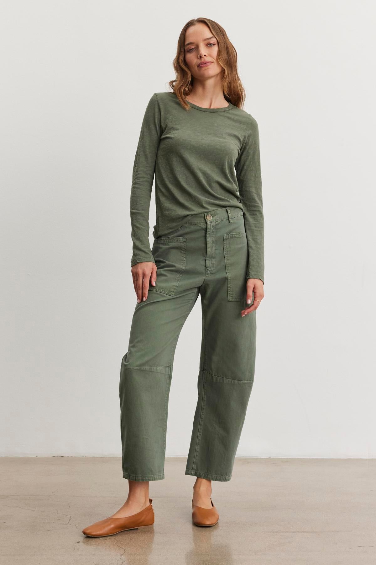   A woman in a green long-sleeve shirt and BRYLIE SANDED TWILL UTILITY PANT by Velvet by Graham & Spencer stands on a beige floor against a white background, completing her utilitarian-inspired look with brown flats. 