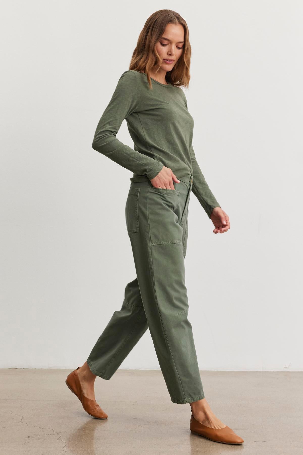 A person in a green long-sleeve top and Velvet by Graham & Spencer's BRYLIE SANDED TWILL UTILITY PANT walks on a wooden floor, wearing brown flat shoes.-38512044540097