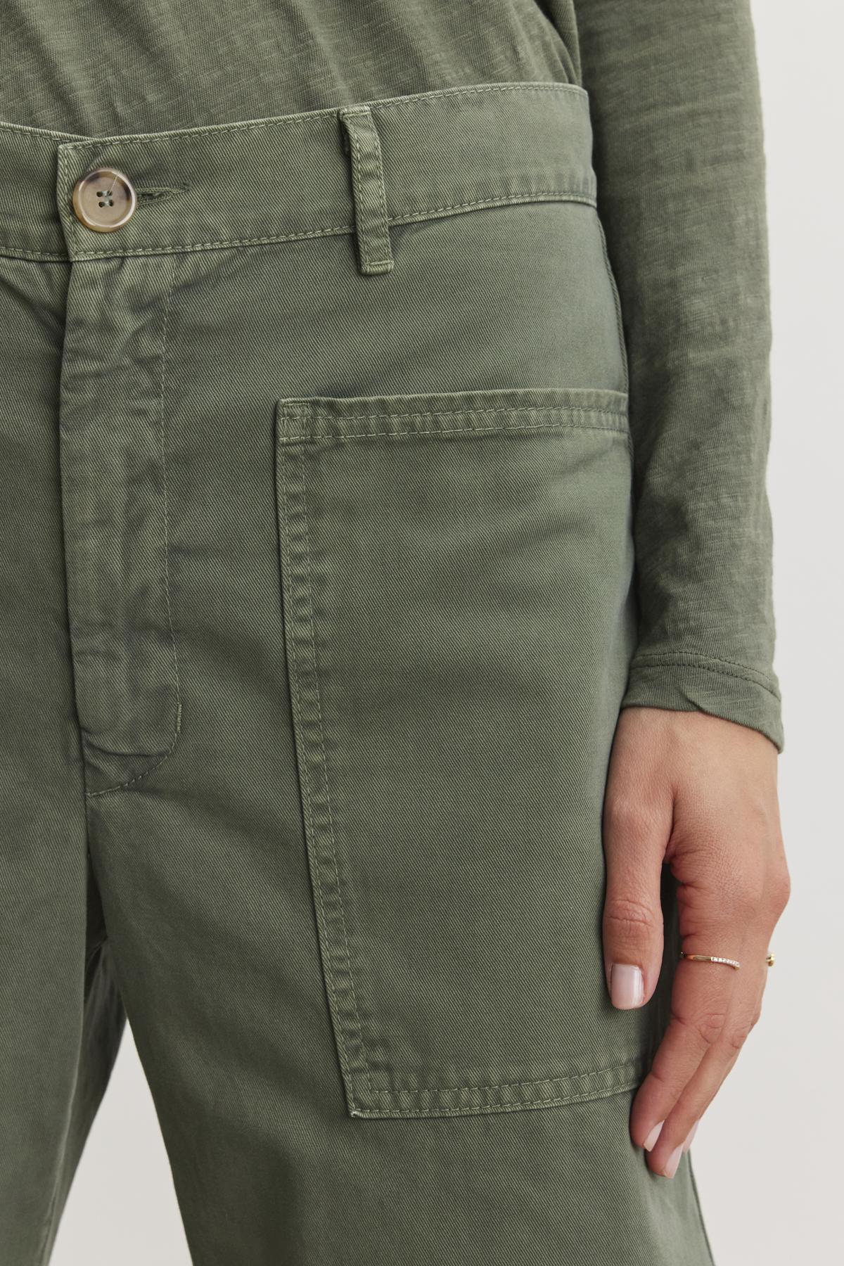   Close-up of a person wearing Velvet by Graham & Spencer's BRYLIE SANDED TWILL UTILITY PANT. The green cotton twill pants feature a large patch pocket, drawing attention as the person's hand rests nearby. 