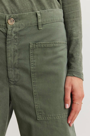 Close-up of a person wearing Velvet by Graham & Spencer's BRYLIE SANDED TWILL UTILITY PANT. The green cotton twill pants feature a large patch pocket, drawing attention as the person's hand rests nearby.