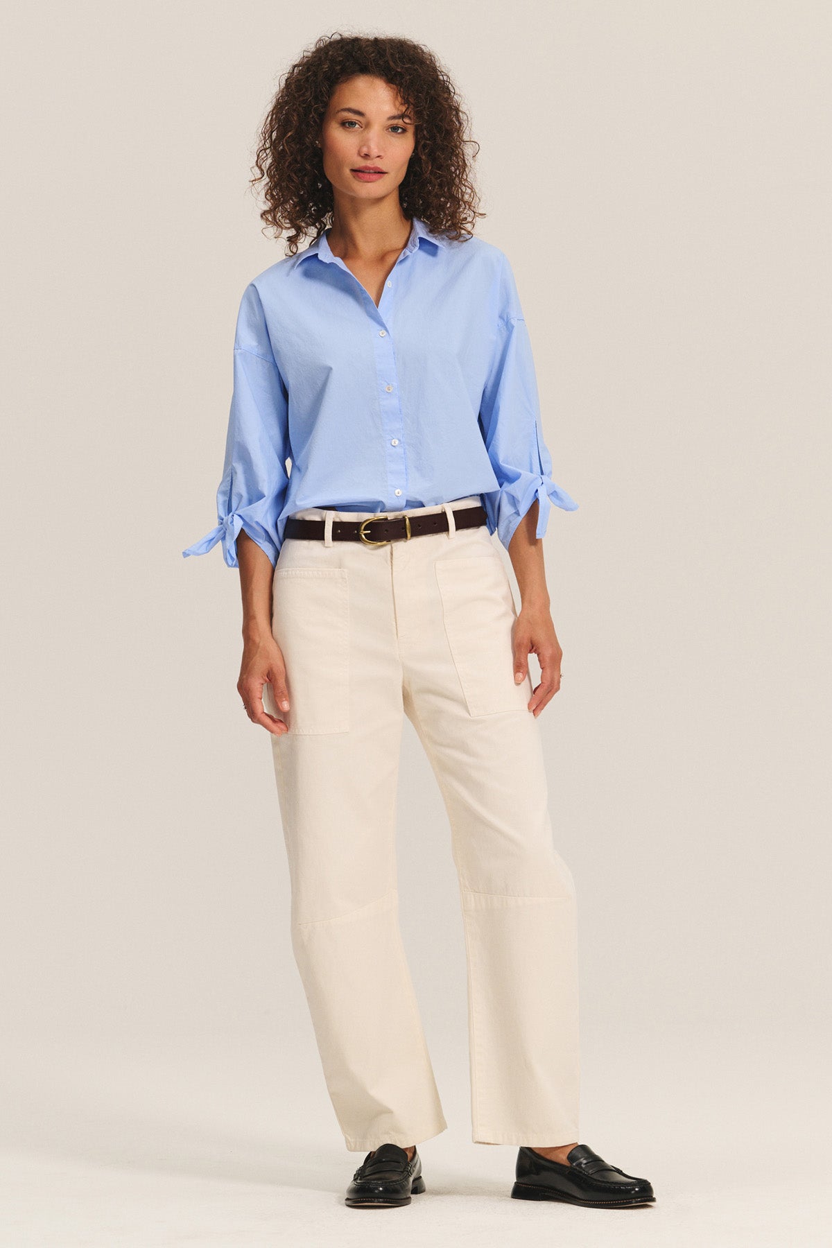 A woman wears the ARLETTE SHIRT by Velvet by Graham & Spencer, a light blue cotton poplin top with a classic collar and sleeve tie details, paired with cream pants, a dark belt, and black loafers against a neutral background.