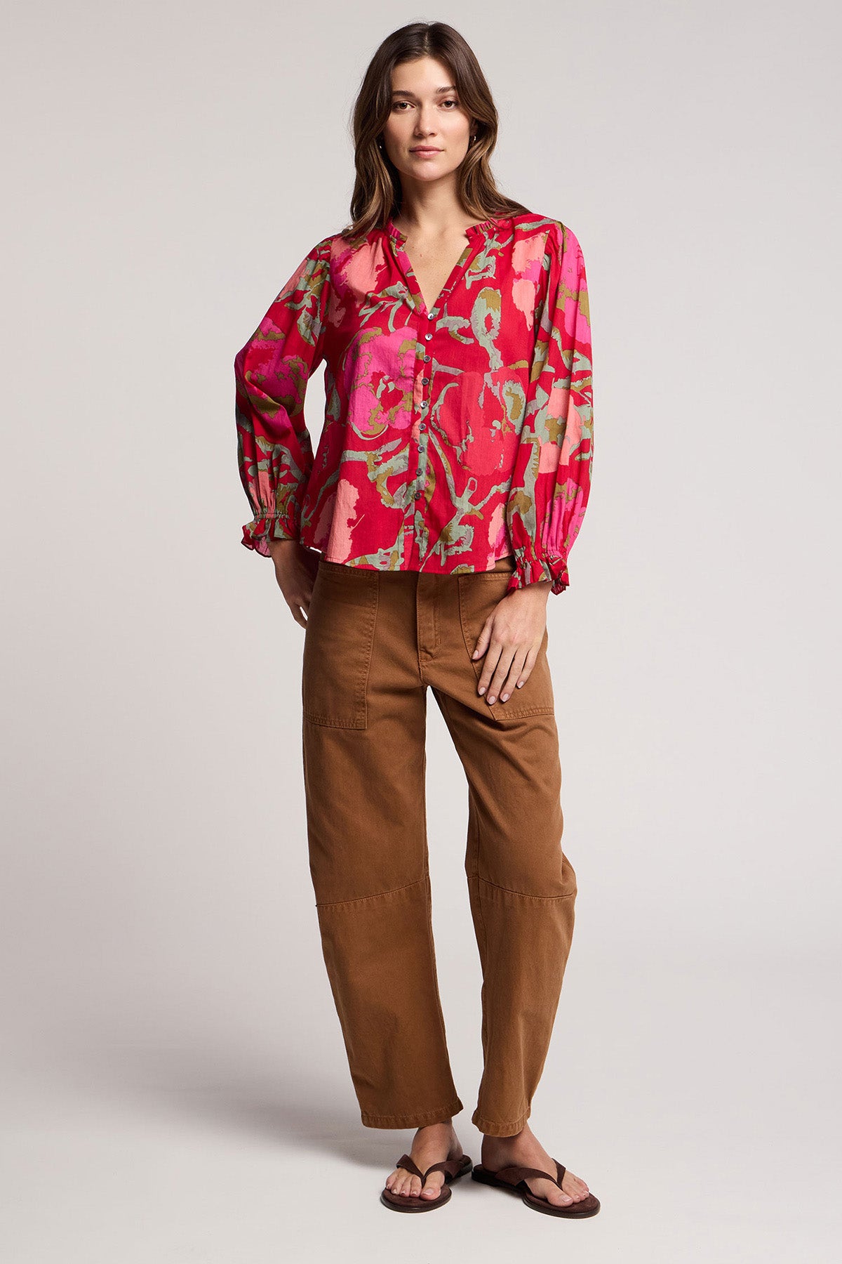 A woman stands against a plain light background wearing the Velvet by Graham & Spencer BRYLIE PANT—cotton twill barrel leg trousers with patch pockets—paired with a bright pink and green patterned blouse and black sandals.