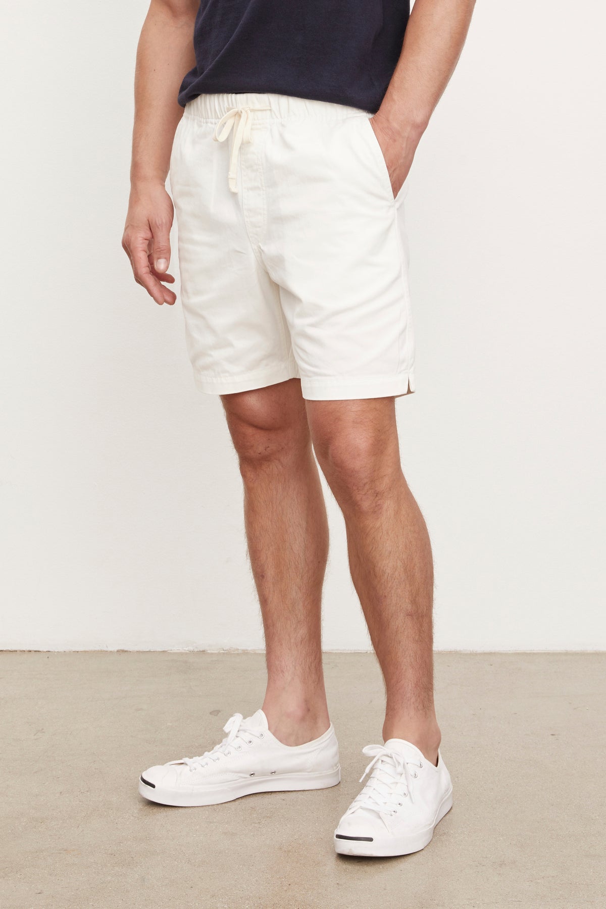   Man wearing a navy blue t-shirt and FIELDER shorts by Velvet by Graham & Spencer with white sneakers, standing in a room with a plain background. Only his torso and legs are visible. 