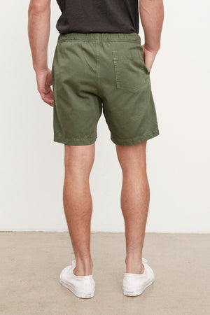 A man wearing Velvet by Graham & Spencer FIELDER SHORT in olive green cotton twill shorts and white sneakers standing with his back to the camera, indoors against a white wall.