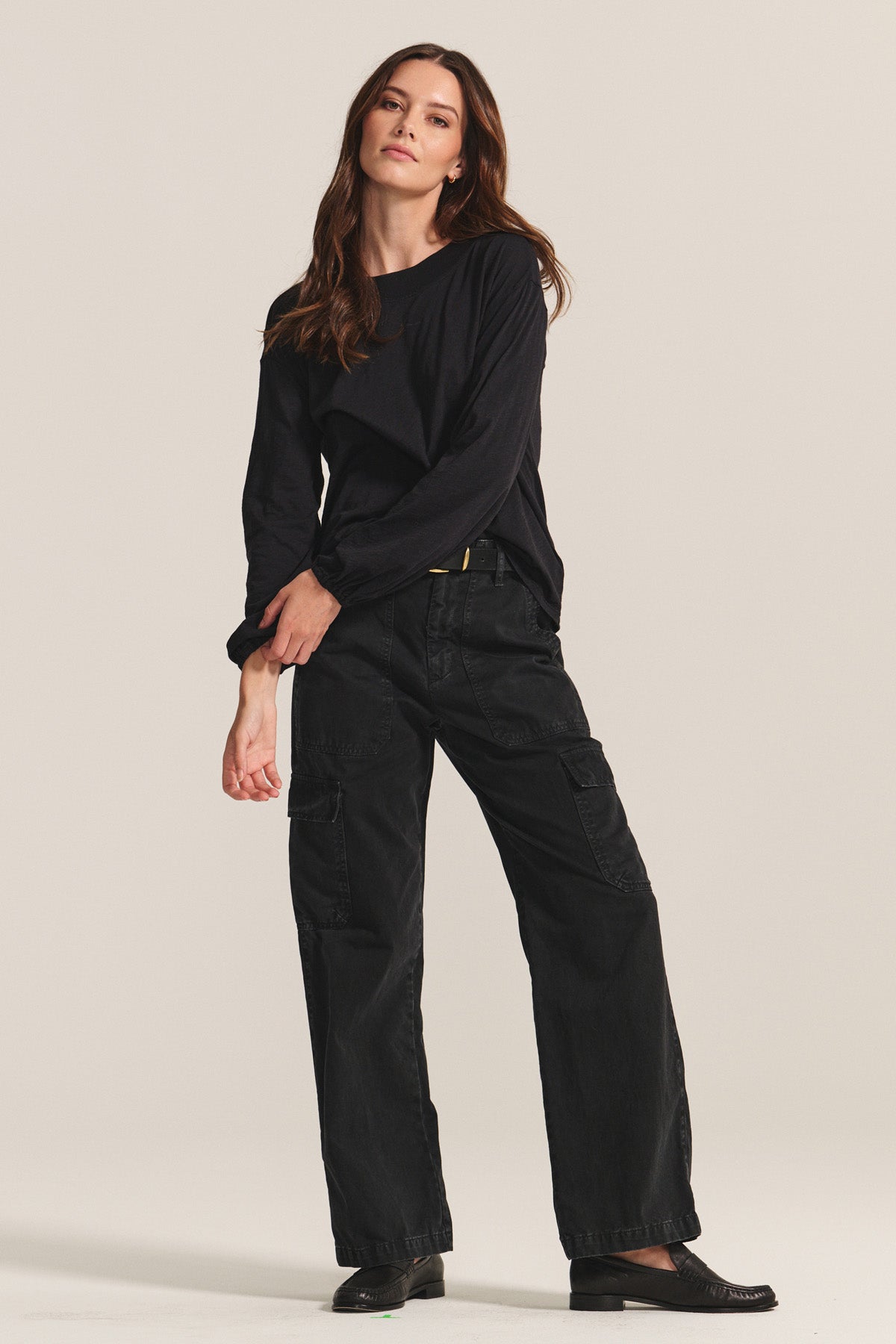 A woman stands against a plain background wearing the JAMINA TEE by Velvet by Graham & Spencer, a loose black cotton slub top, paired with black cargo pants and black shoes.-40688692756673